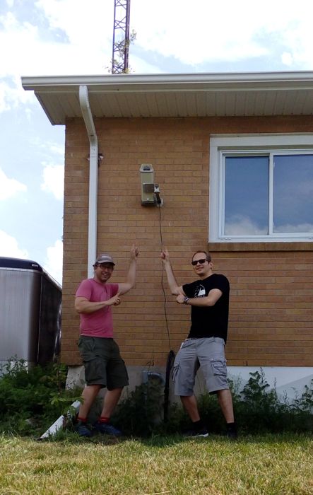 Two men pointing at a small camera mounted on the brick wall of a building
