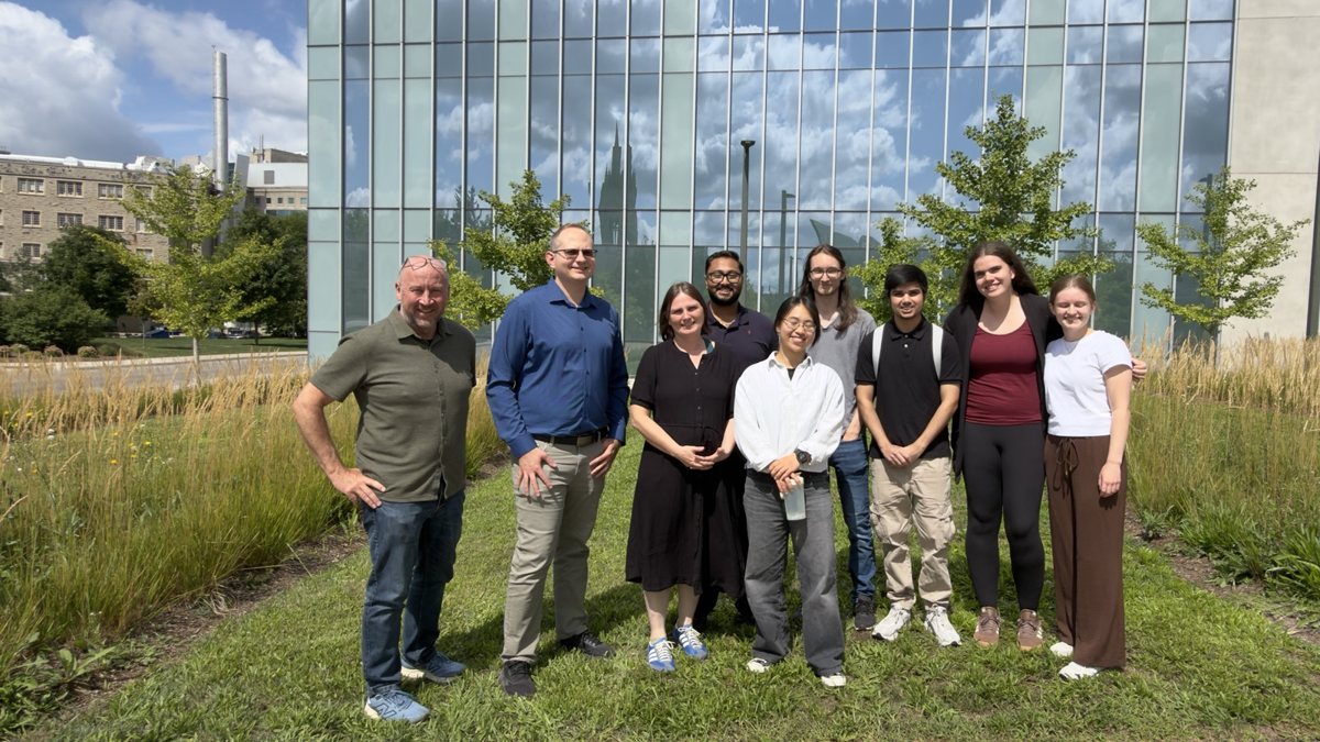 Ten members of the contrail team standing on the grass in front of a glass building on Western's campus that reflects a blue sky with clouds